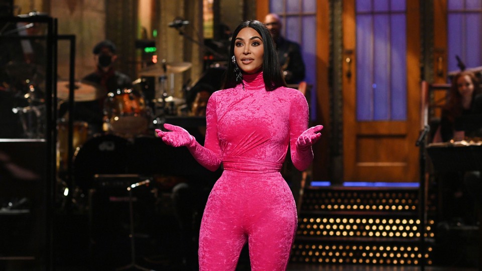 Kim Kardashian standing palms up on the Saturday Night Live stage, wearing a tight pink bodysuit that covers her entire body, including her hands.
