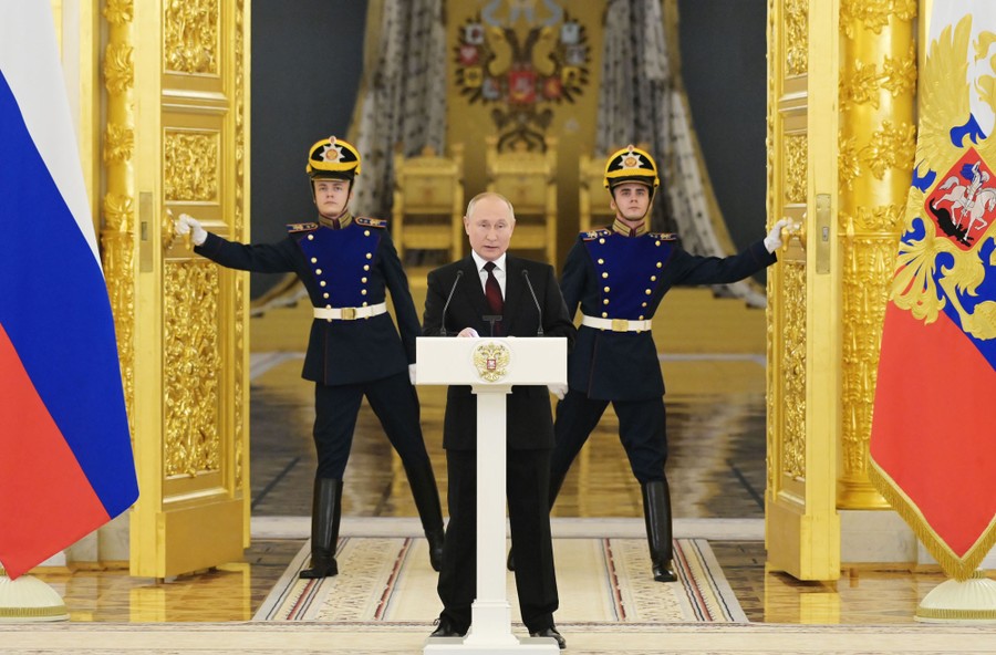 Two soldiers in ceremonial gear operate large ornate doors behind Vladimir Putin as he speaks at a lectern.