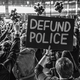 Fotografía en blanco y negro de una multitud que protesta con los puños en alto y un cartel que dice, “DESFINANCIAR LA POLICÍA”.