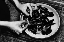 A black and white photo of someone cleaning mussels, with someone else's shoes in the photo, in black strappy sandals