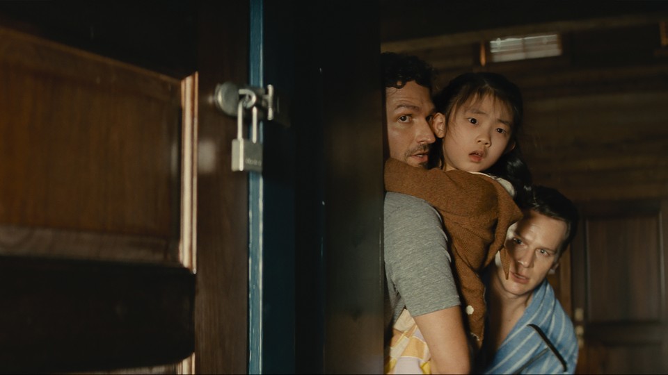 Jonathan Groff, Ben Aldridge, and Kristen Cui hide behind a cabin door in "Knock at the Cabin."