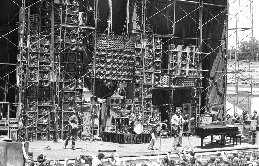 The Grateful Dead perform on a stage in front of an enormous wall of stacked speakers.