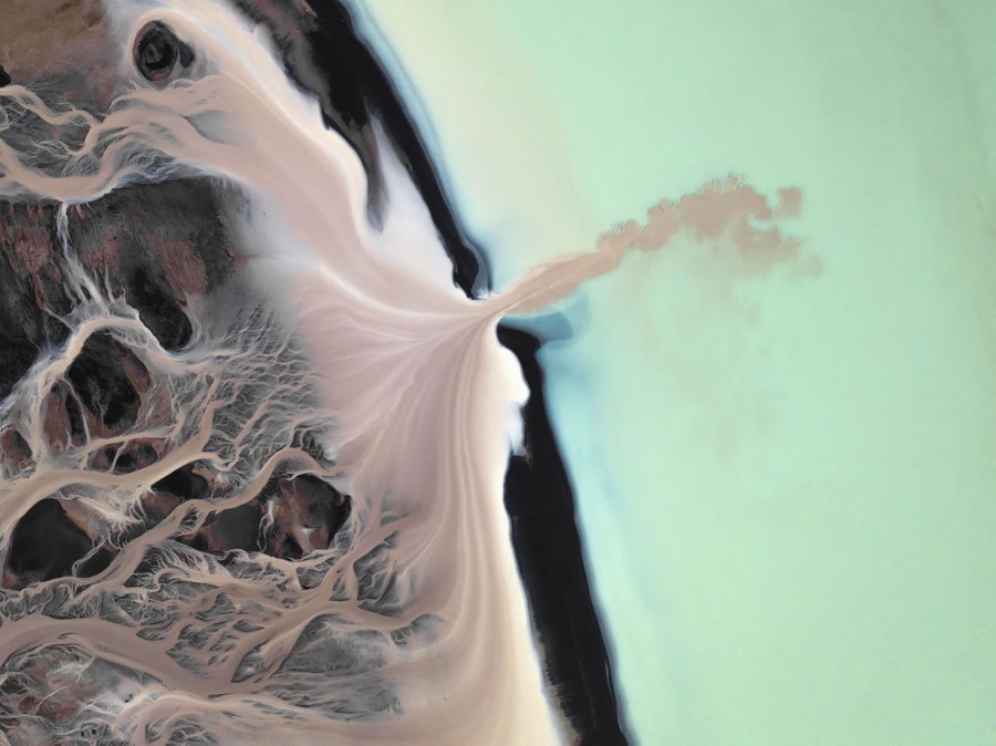 A top-down aerial view of tan-colored water flowing from a many-branched riverbed into a light-green body of water