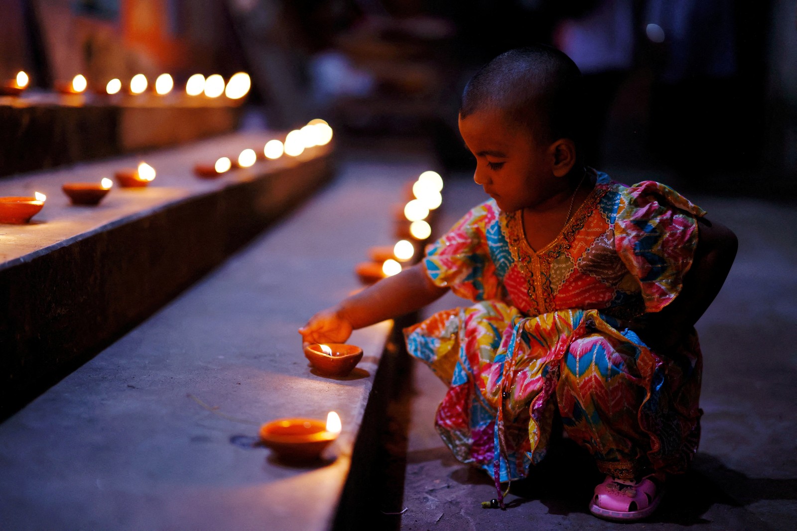 A child kneels down to touch one of many small oil lamps laid out on temple steps.