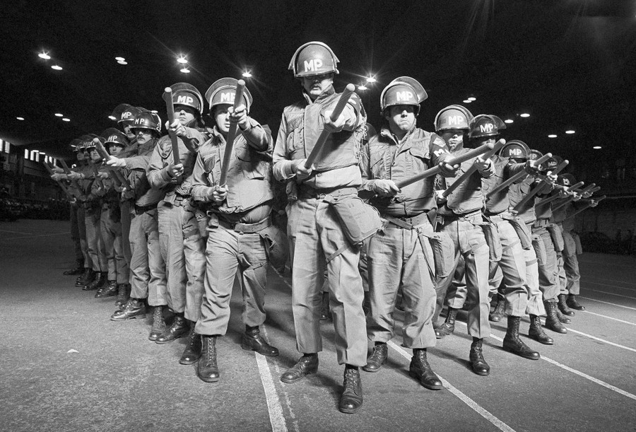A small group of national guardsmen stand in a V-formation, holding out their batons.