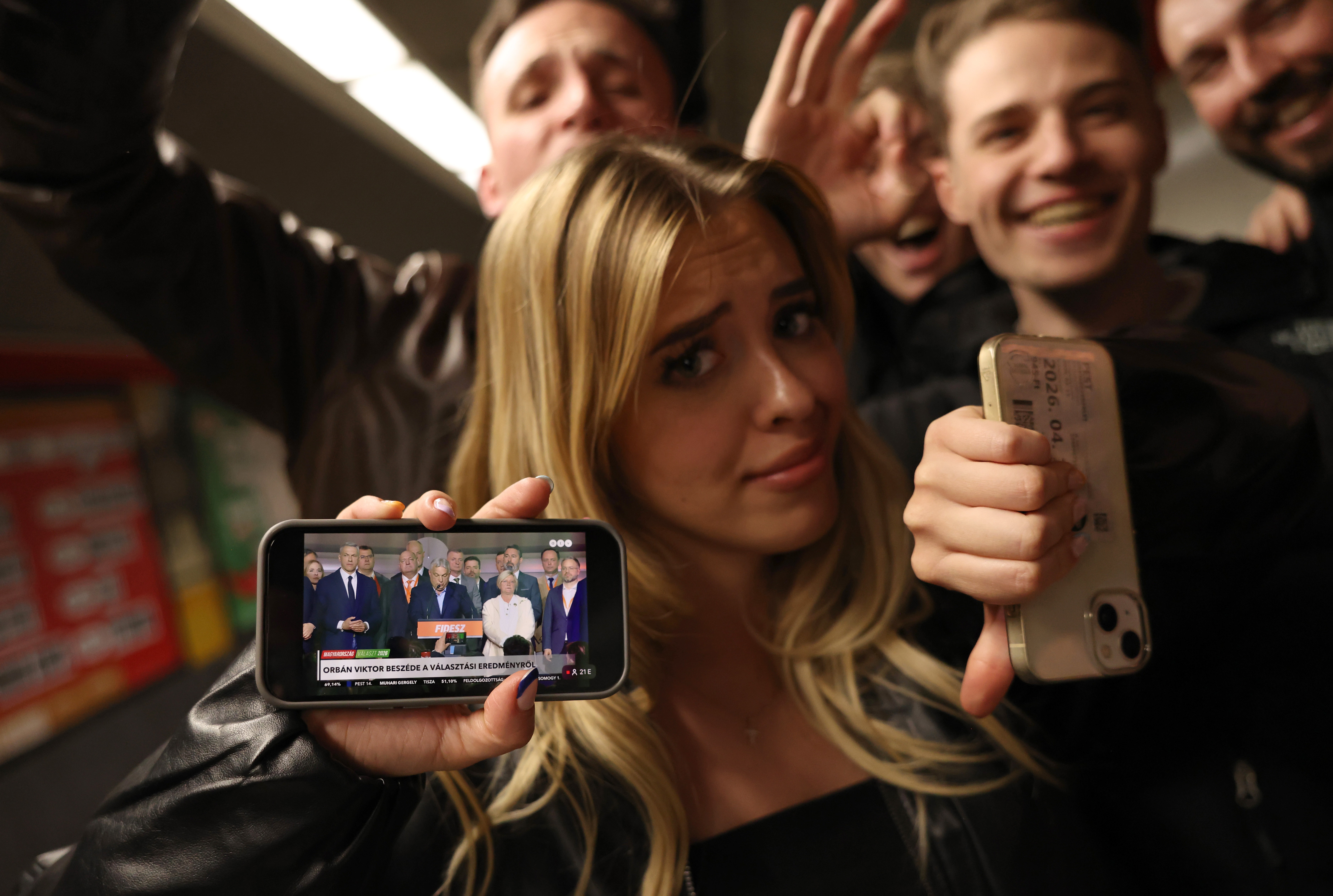 One of a group of celebrants in an underground metro station gives a thumbs-down while showing live video of a concession speech on their phone.