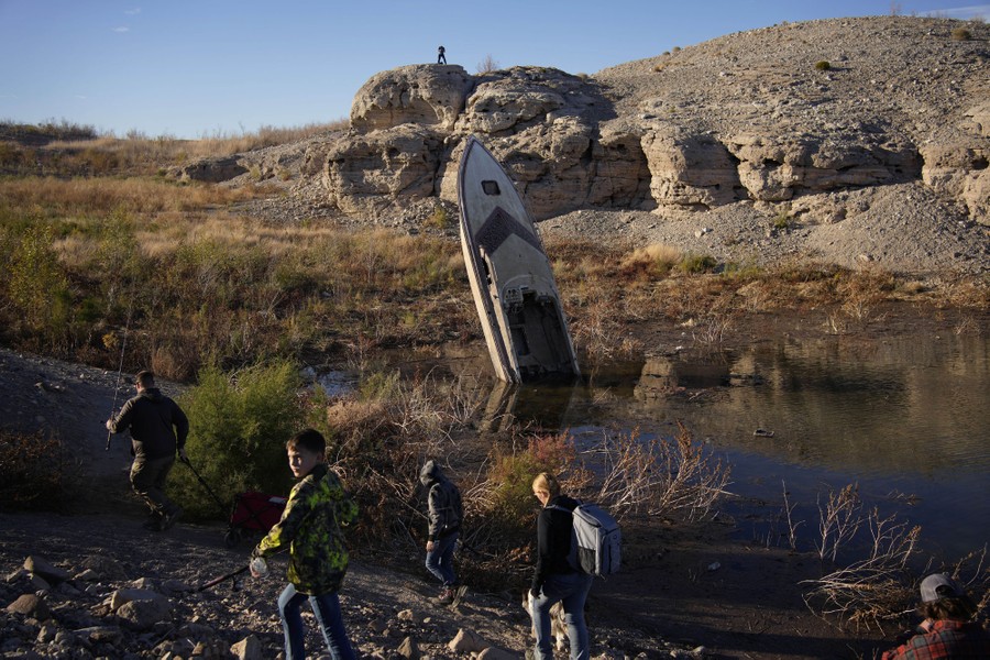 People walk past the old wreck of a boat left standing nose-up in the drying mud of an emptied reservoir.
