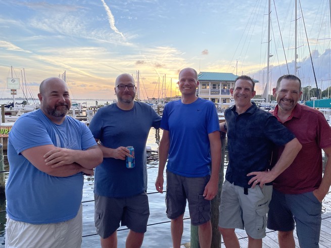 Five men smile in front of a sunset over a harbor