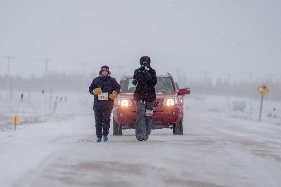 Two people dressed in cold-weather clothing run on a road, followed closely by a car, during a snowstorm.