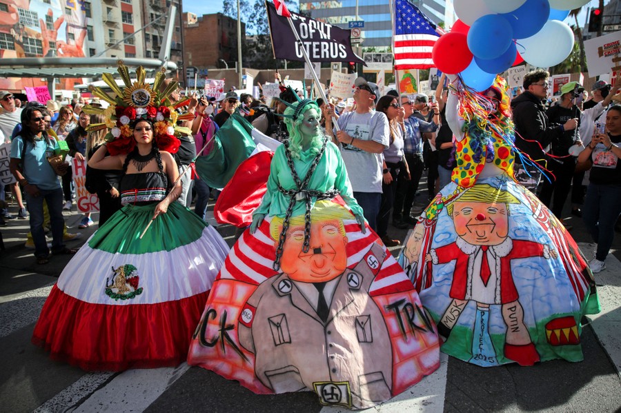 Several protesters wear elaborate costumes depicting a Mexican flag and unflattering caricatures of Donald Trump.