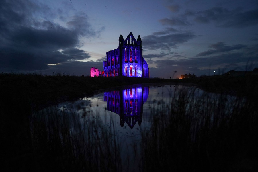 An abandoned abbey is seen reflected in a pond at night, lit red and purple.