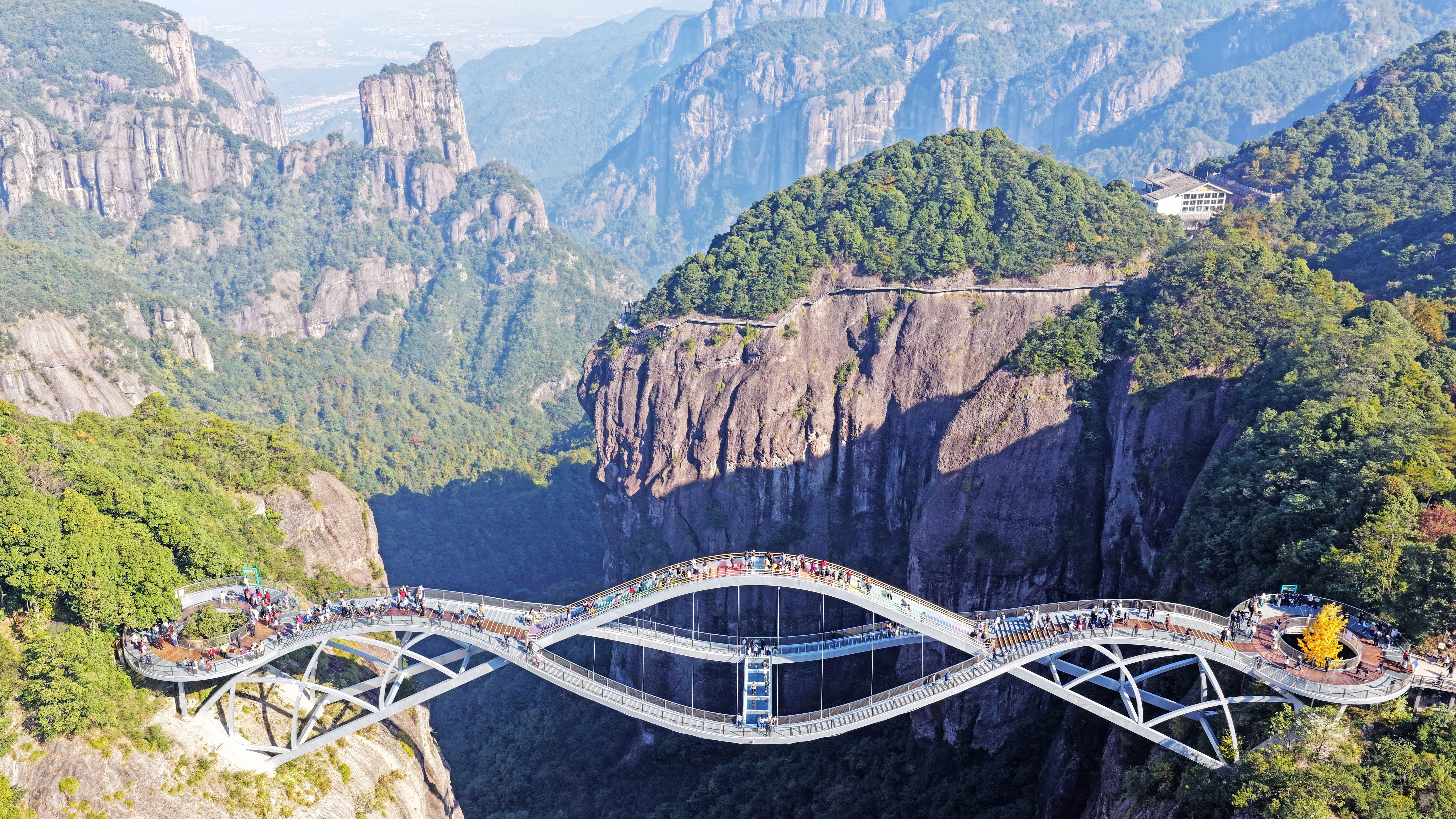Tourists walk on an undulating glass-bottomed bridge over a steep valley.