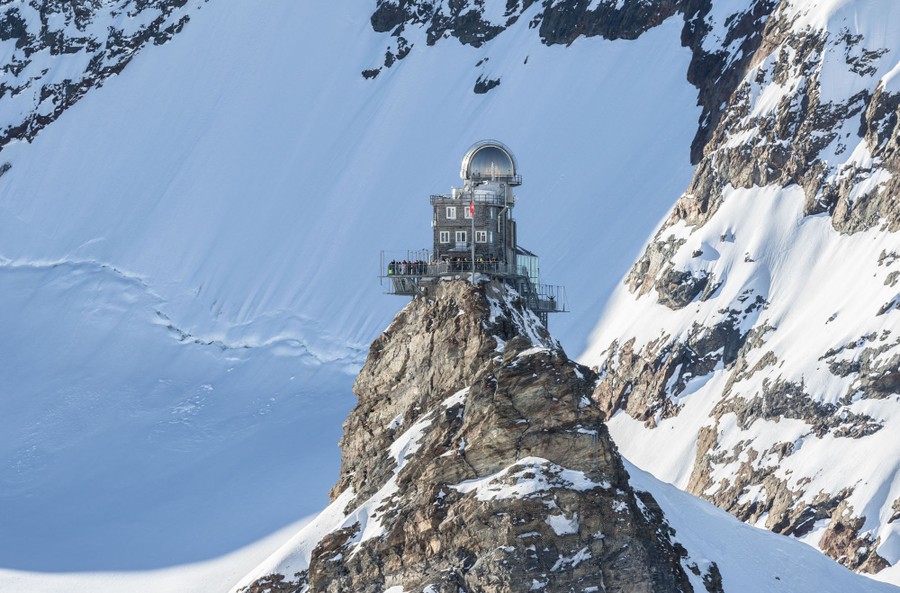A small observation building stands on a peak among larger mountains.