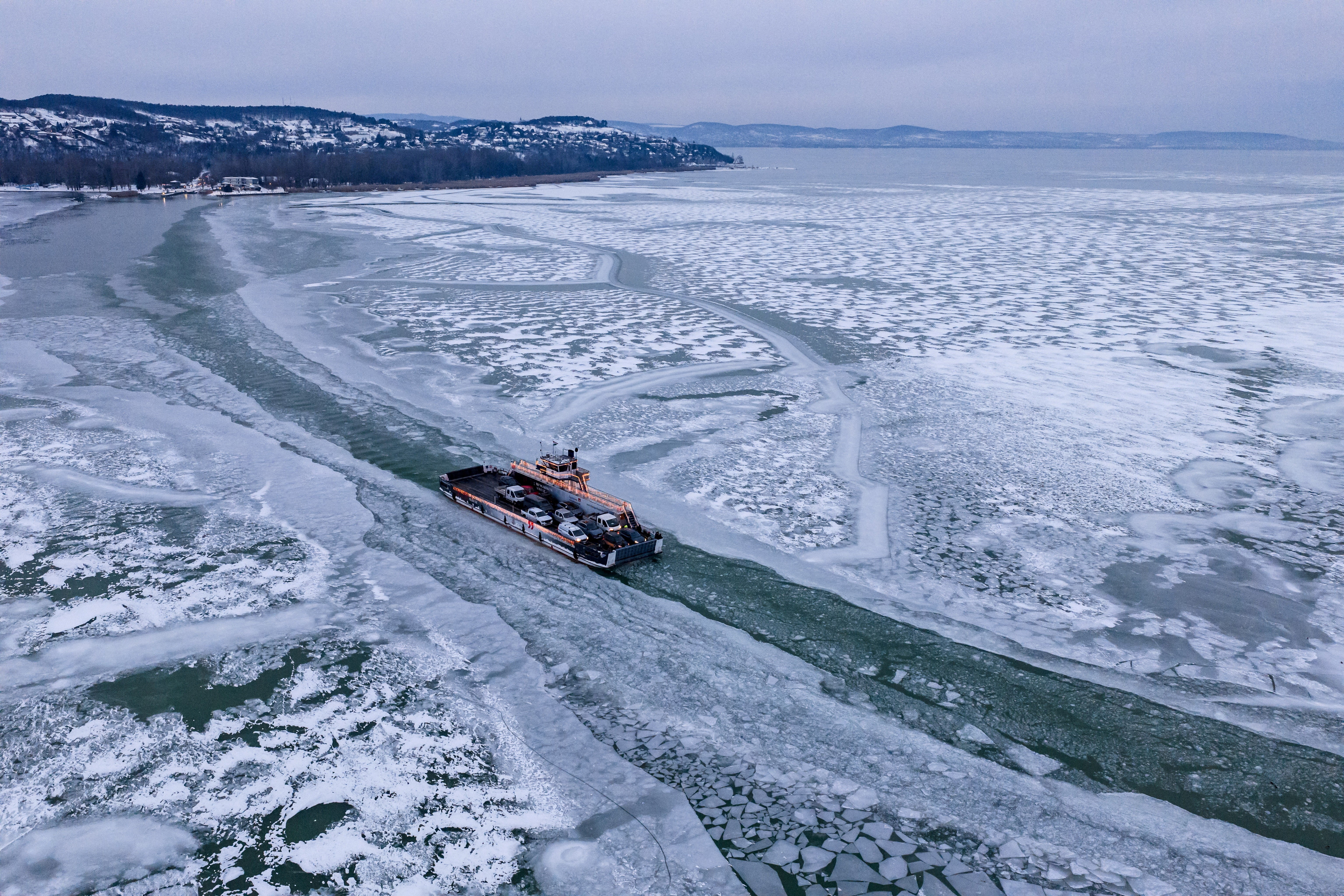 A drone view shows a ferry navigating a channel through a frozen lake.
