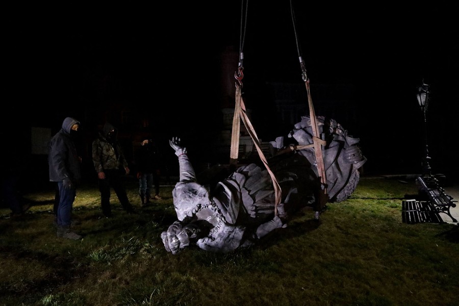 A large bronze statue is lifted from a park with straps, lying on its side.