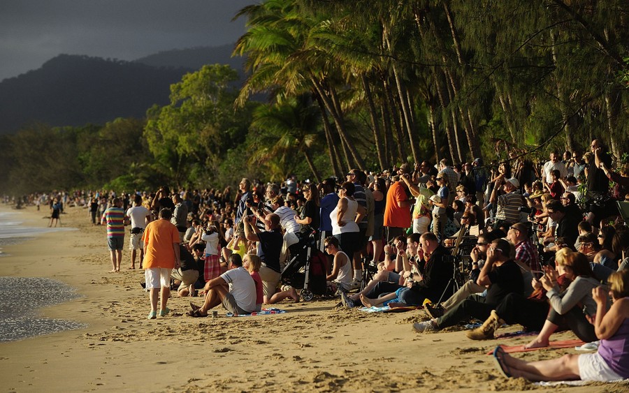 Hundreds of people stand and sit along a beach, looking toward the sun.