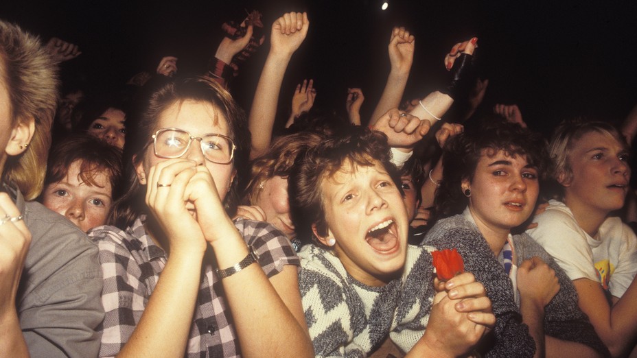 A crowd of pop music fans cheering at a stage. 