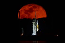 A rocket stands on a launch pad, lit at night, seen from a distance, with a red-colored full moon rising in the background.