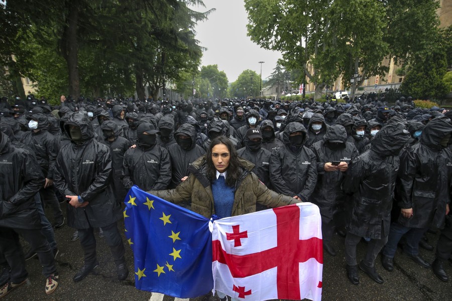 A woman stands in front of a large group of masked riot police officers. She holds a Georgian national flag and an EU flag tied together.