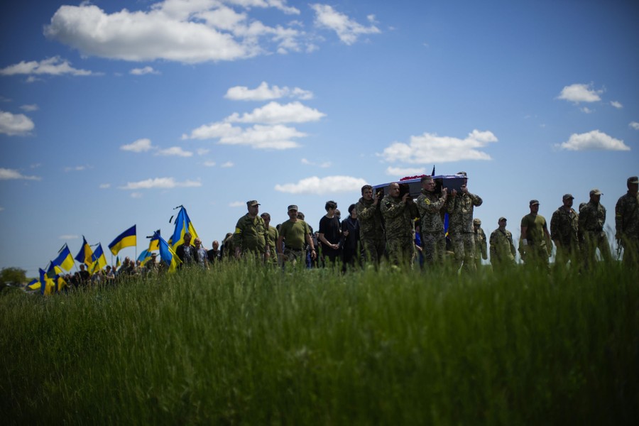 A line of people follow pallbearers carrying a coffin past a grassy area.