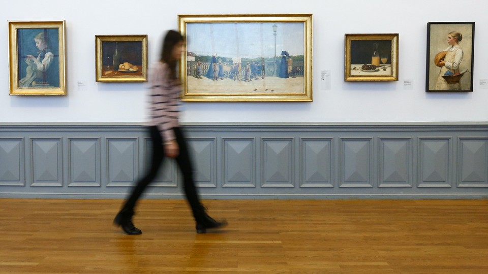 A woman, who is blurry, walks past a series of paintings in a museum.