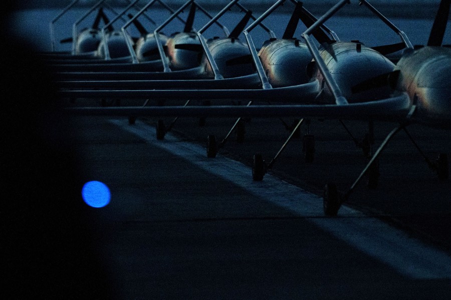 Eight airplane-shaped attack drones sit, lined up, on a runway.