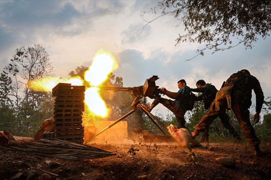 Several people make an effort to fire a heavy machine gun that is mounted on a tripod, lit by the bright flash from its muzzle.