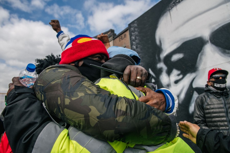 Several people embrace in front of a mural of George Floyd.