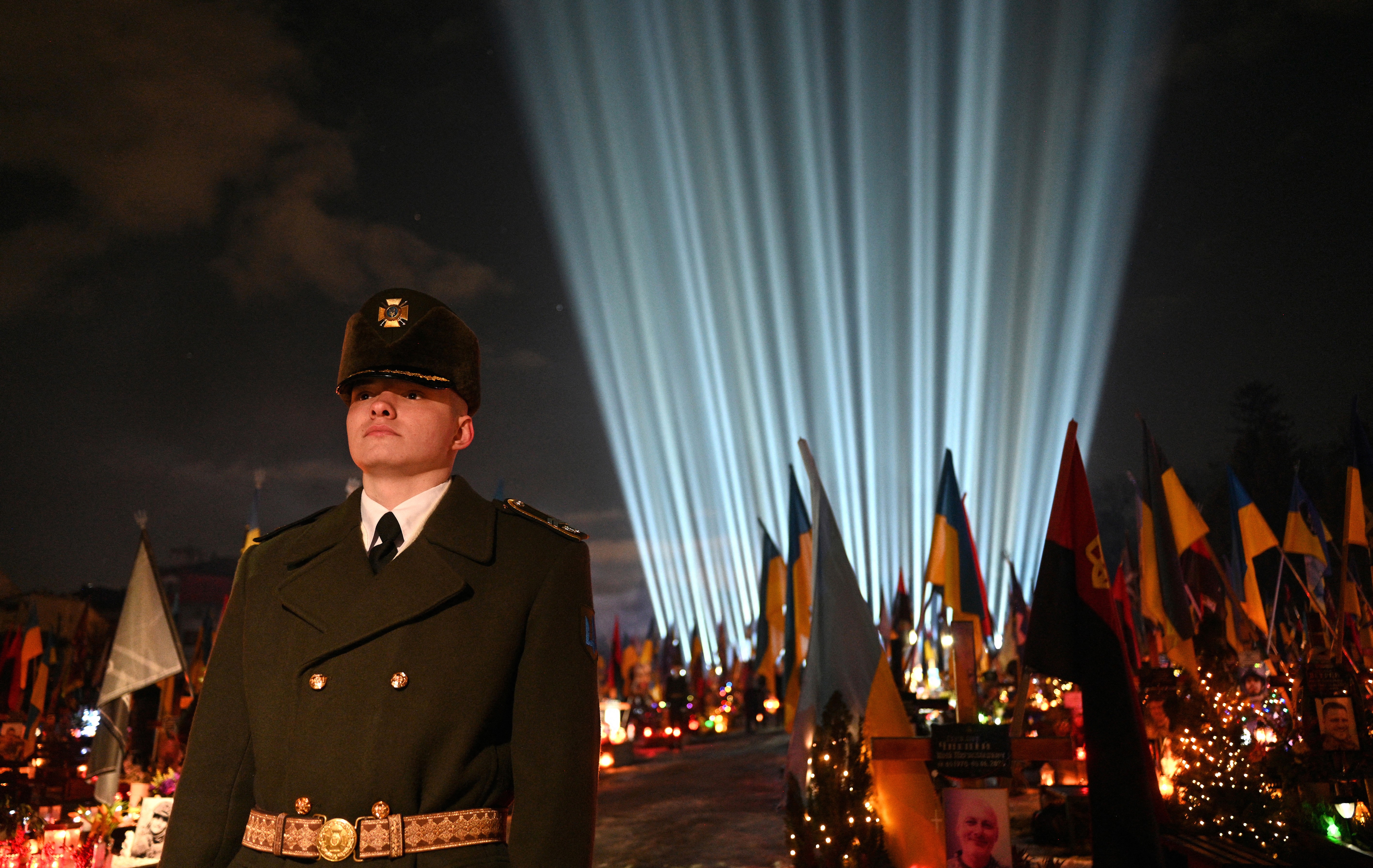 A soldier in dress uniform stands guard in front decorated military gravestones and a light display.