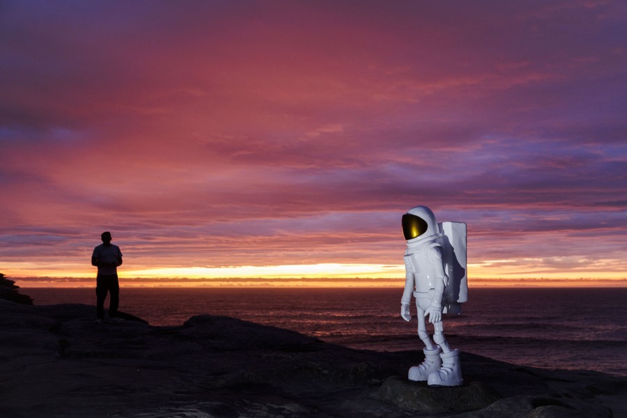 A person stands near a sculpture of an astronaut, along a shoreline, just before dawn.