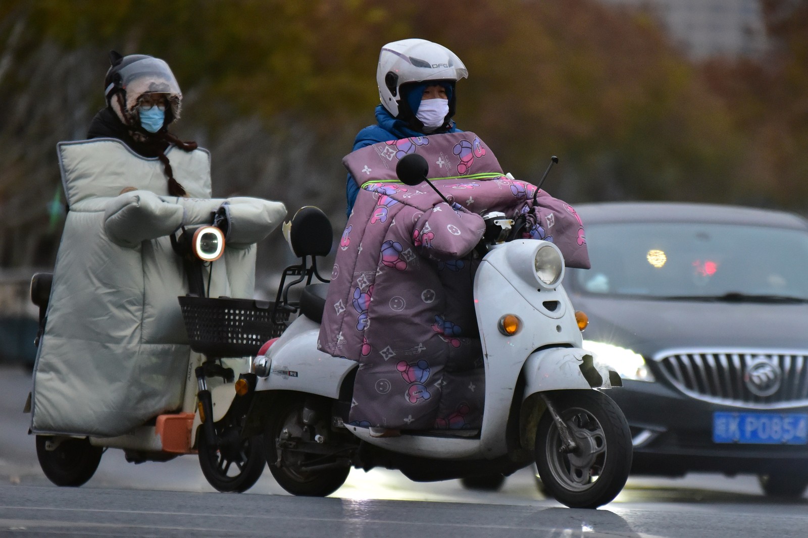 Two people ride on scooters, with custom-made blankets wrapped over their hands, handlebars, and the front of their body.