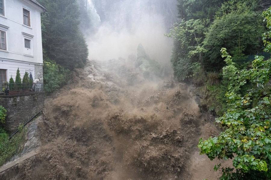 Muddy water splashes down on rocks at a flooded waterfall, beside a building.
