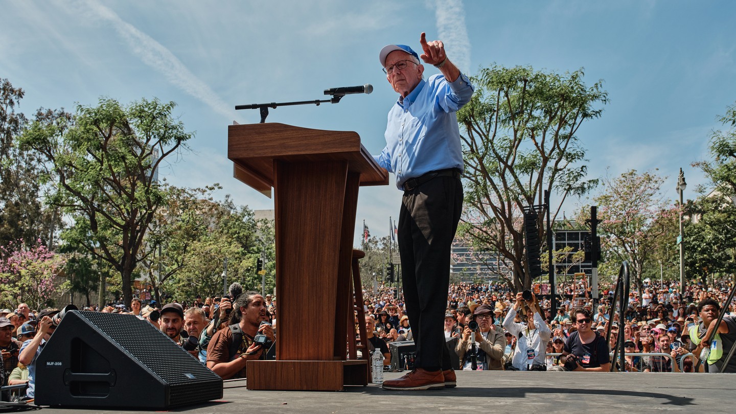 Bernie Sanders stands on a stage at a podium and points out