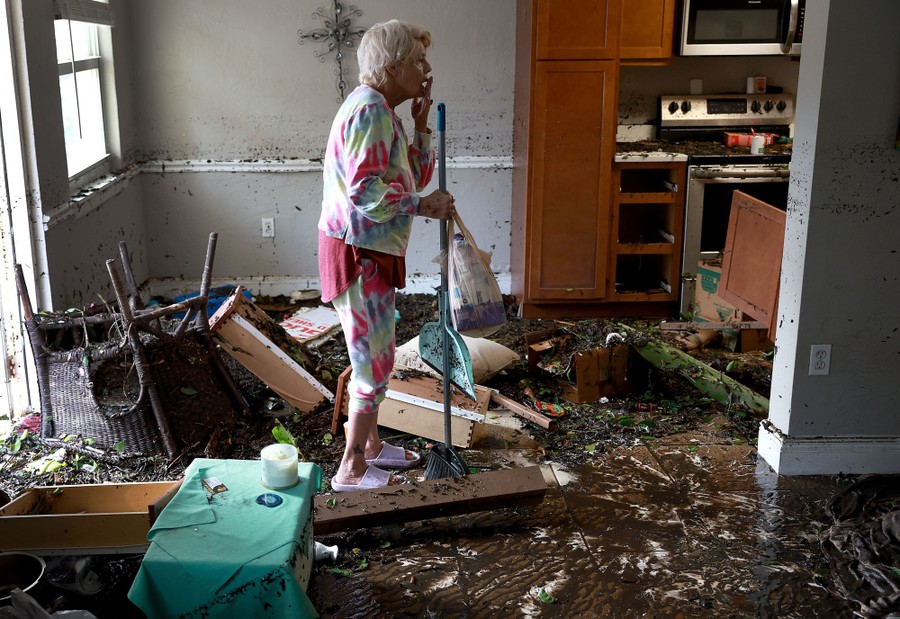 A person holding a broom stands in the middle of their flood-damaged apartment.