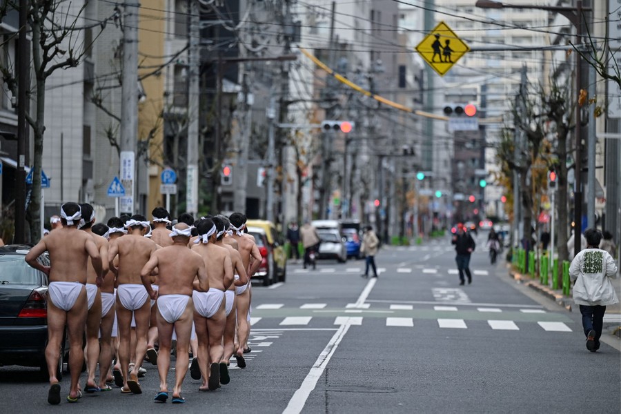 A group of men wearing nothing but traditional loincloths jog together in a city street.