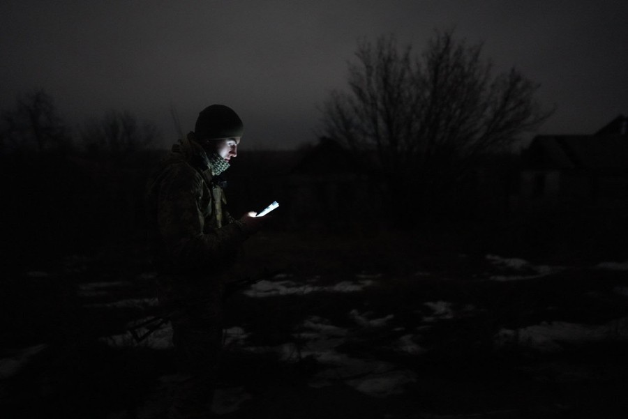 A soldier looks at the screen of a handheld device, in a field, at night.