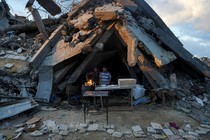 Two men stand at a table next to an open flame used for baking, with a huge mound of rubble rising up behind them.