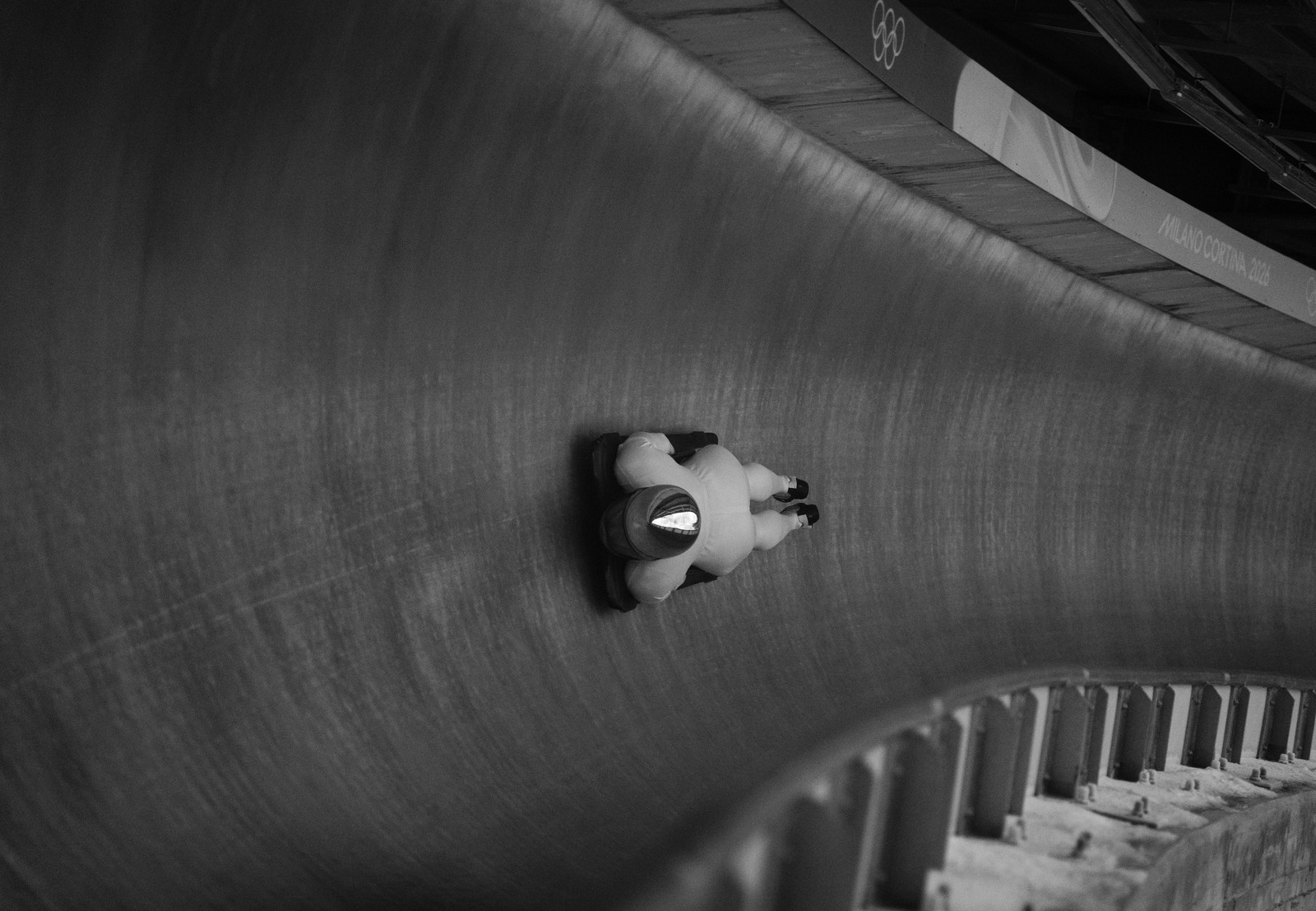 An infrared view of a skeleton racer on their sled, pinned to the side of a steep-walled ice track as they fly past.