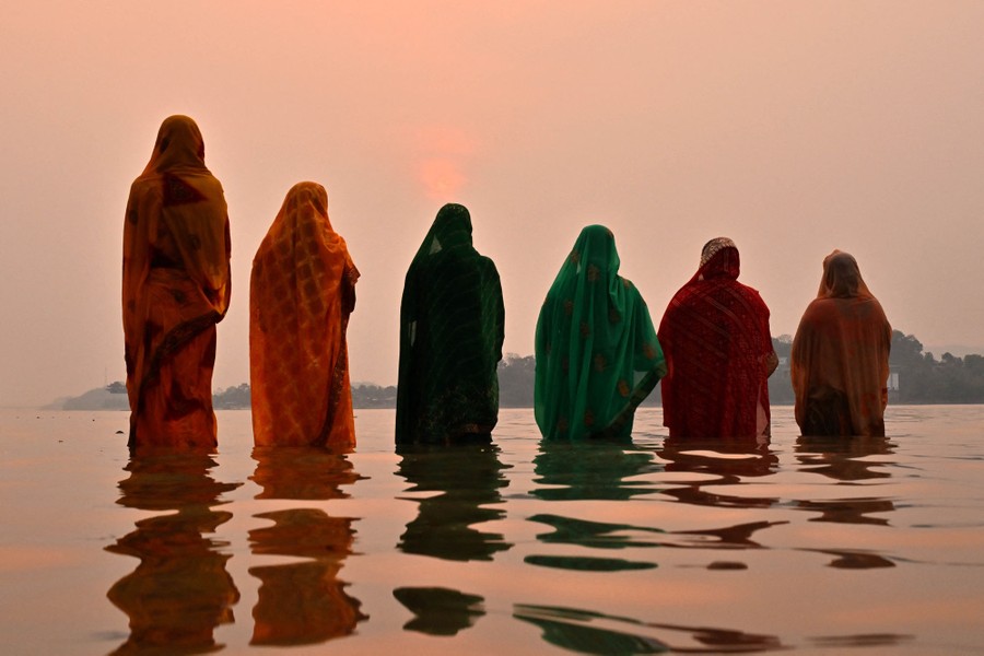 Six women in colorful garments stand in shallow water while praying.