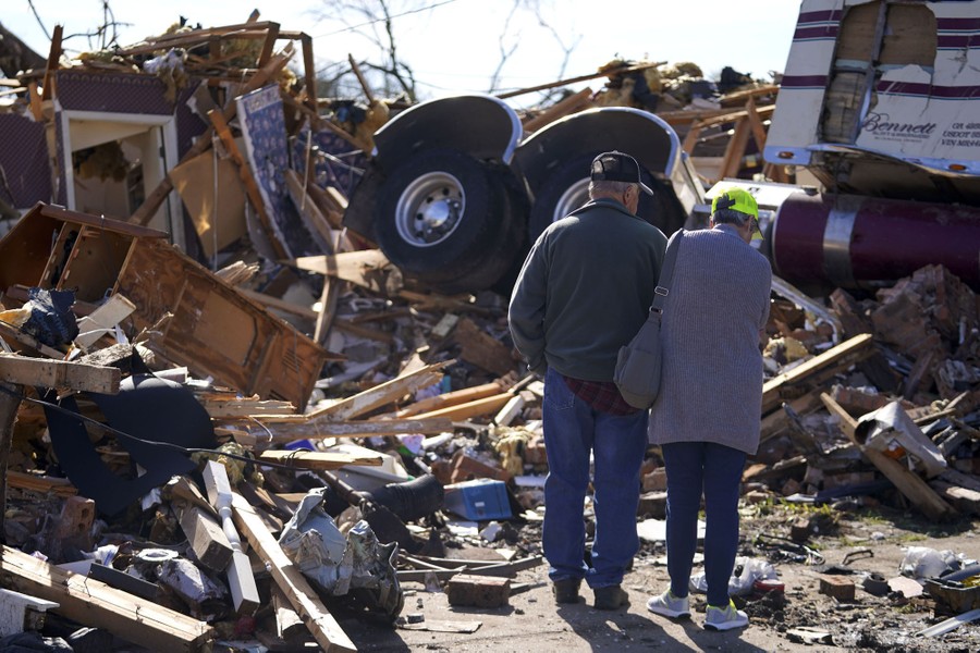 A couple stands close together, surveying extensive damage from a tornado, including a tractor unit of a semi-trailer truck atop a pile of debris.