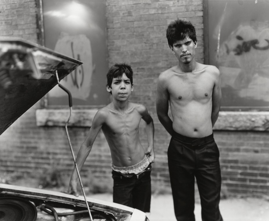 black-and-white photo of younger and older brothers standing shirtless next to car's engine with propped-open hood, with brick wall and bricked-in, graffitied windows in background