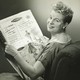 A black-and-white image of a woman reading a 1950s newspaper