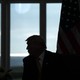 A color photograph of Donald Trump seen in silhouette against tall windows.