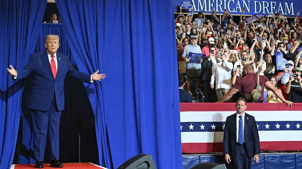 Donald Trump stands on a stage with blue curtains, hands outstretched
