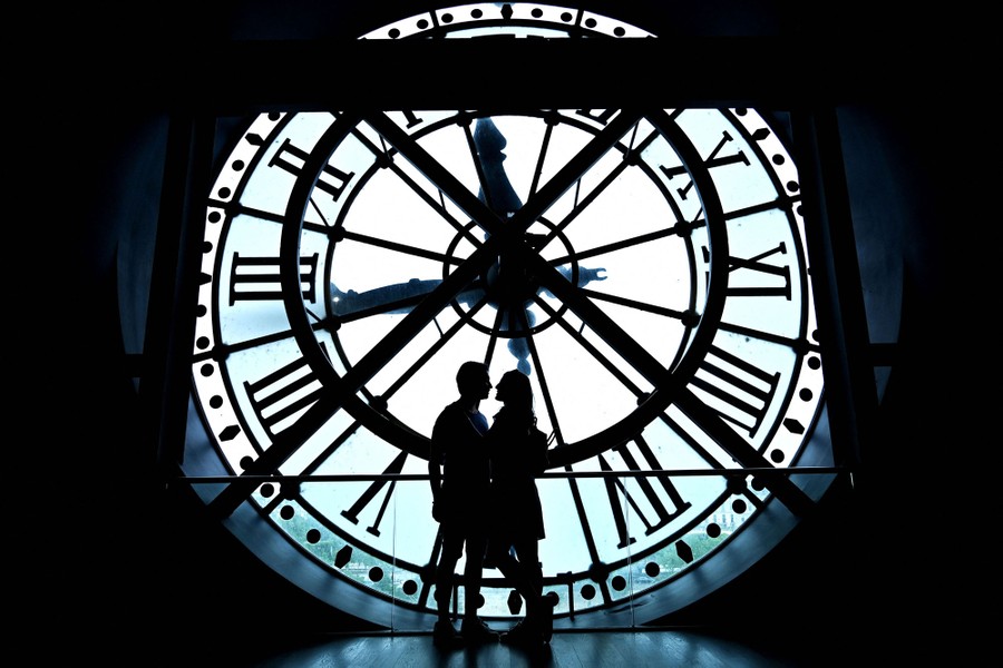 A couple poses for a photo near a giant clockface, seen in silhouette.