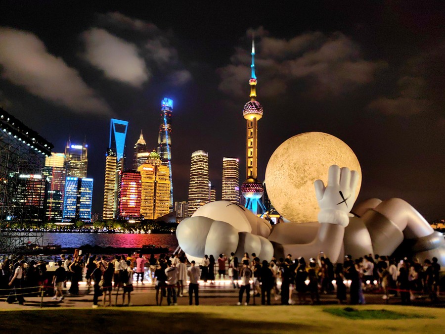 A giant inflatable art installation of a figure lying on its back, holding a moon, with the Shanghai skyline in the background