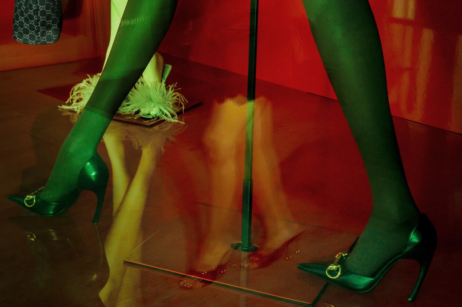 color photo of retail window with green-tinted mannequin legs in heels, saturated red background, and reflection of person shopping