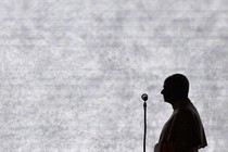 Photograph of Pope Leo’s black silhouette from the side as he speaks in front of a microphone.