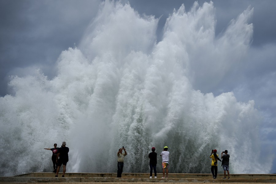 Half a dozen people stand beside a seawall, taking photographs as large waves splash in the air.