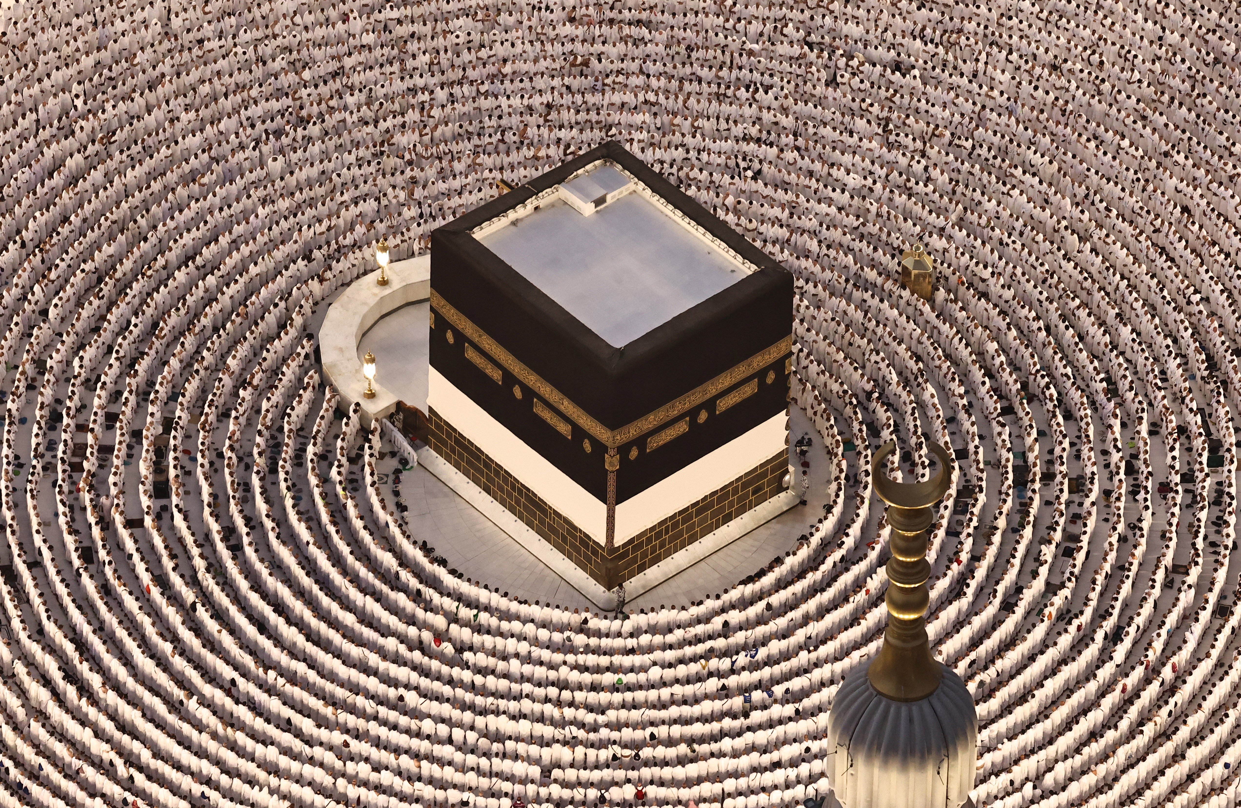 Many Muslim pilgrims dressed in white kneel in prayer, arranged in concentric circles around a tall cube-shaped shrine.
